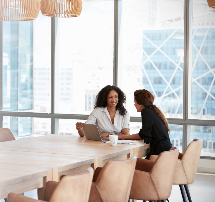 Two women sitting at the corner of a large table in front of a sky scraper window